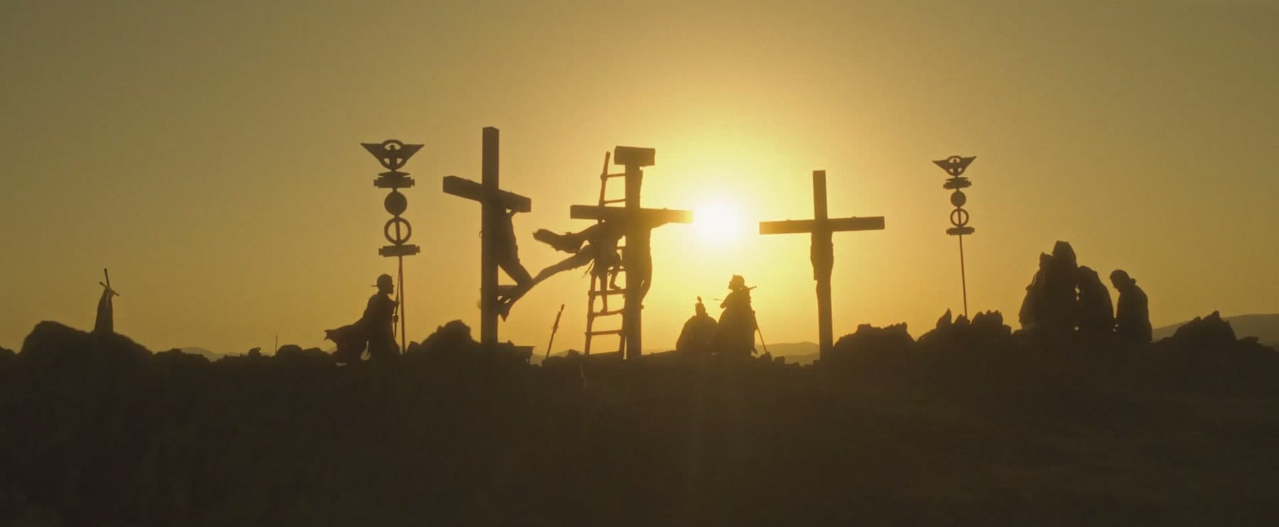 Three crosses silhouetted on a hilltop against a golden sunset, soldiers and mourners gathered below — a still from the film